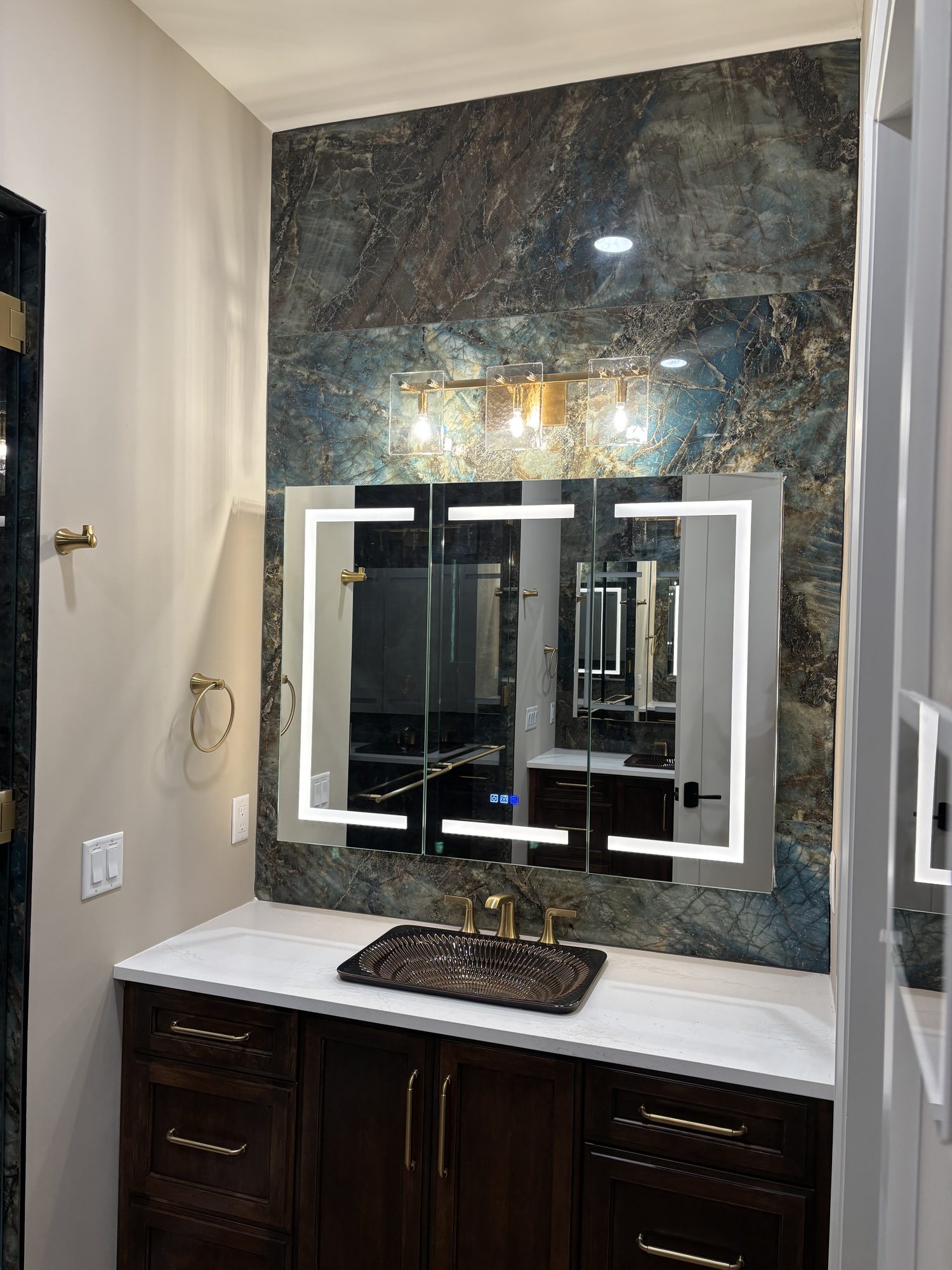 Backlit LED mirrors with sconce vanity lighting over quartzite feature wall in Tampa custom home bathroom by Dim and Dimmer Electric